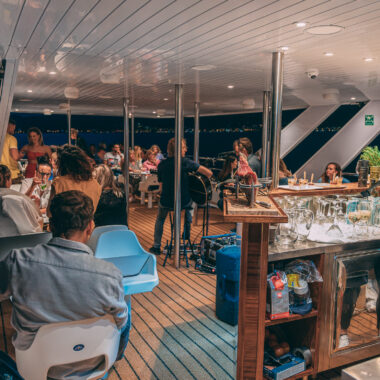 Barmen serving drinks, musicians playing, a guest recording the scene with a mobile phone, and guests enjoying a sunset cruise